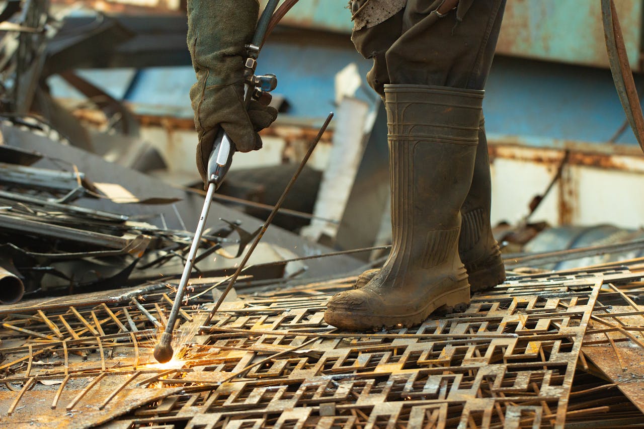 A worker uses a cutting torch on scrap metal in a yard, with sparks flying around.