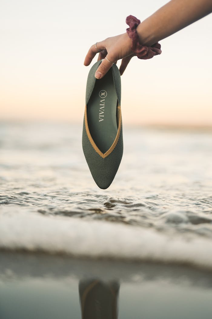 A green shoe held by a hand near ocean waves during sunset, creating a serene scene.